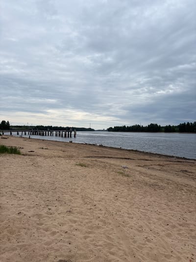 The image shows a sandy beach in Nevskaya Dubrovka, Russia, with a wide body of water in the background. A dilapidated pier, with wooden pilings and concrete structures, extends out into the water from the left side of the frame. Trees line the opposite shore, and a transmission tower is visible in the distance. The sky is overcast with gray clouds. The sand in the foreground is textured with ripples and scattered debris, including what appears to be a pair of white shoes and some plastic. The water is calm and reflects the cloudy sky. The overall scene suggests a somewhat neglected, natural waterfront environment. There are no people visible in the image.