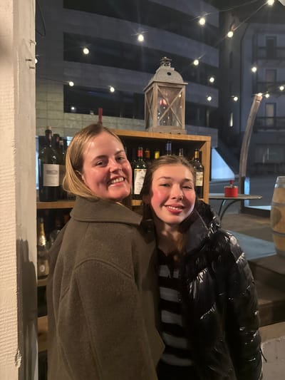 This nighttime image, captured in Sankt Moritz, Switzerland, depicts two smiling adult females in an outdoor or semi-enclosed setting. The woman on the left, with blonde hair, wears a dark olive-colored overcoat and smiles directly at the camera. The woman on the right, with brown hair, wears a black shiny puffer jacket over a striped shirt and also smiles at the camera. Behind them, a wooden display unit holds numerous dark wine bottles and a rustic wooden and metal lantern. String lights are suspended overhead, providing illumination. To the right, a dark round table with a red circular ashtray or container and a partial wooden barrel are visible. The background features dark multi-story buildings with illuminated windows. A textured white column borders the left side of the frame.