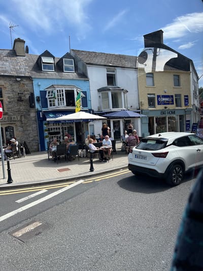 This media file captures a street scene in Donegal, Ireland, under a bright, sunny sky with scattered clouds, suggesting a pleasant daytime. The foreground is dominated by a paved sidewalk lined with an asphalt road marked with double yellow and white lines. Several outdoor seating areas for cafes or restaurants are set up on the sidewalk, with patrons enjoying refreshments.

The buildings lining the street are a mix of architectural styles. One building features a light-colored facade with windows and signs, including "ESKE HOME" and "O'DONNELL'S Music Shop," indicating retail establishments. Another building, painted a light blue, has a prominent sign for "The Tea Room & Restaurant" with "Chef Owner" and "Old Cove" also visible. Green and yellow flags are displayed on this building. The stone facade of another building is partially visible at the bottom left.

A white car with an Irish license plate "261-D-29787" is parked on the right side of the road. People of various ages are seated at the outdoor tables, engaged in conversation and enjoying the weather. Some individuals are partially obscured by umbrellas. The atmosphere appears relaxed and social, with people taking advantage of the good weather to dine outdoors. The overall scene depicts a typical day in a charming Irish town, with a focus on outdoor dining and local businesses.