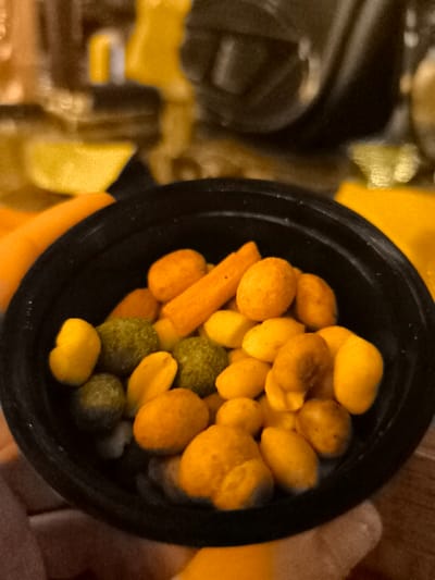 A close-up shot captures a black bowl filled with an assortment of colorful snacks. The primary contents include numerous orange-coated nuts or peanuts, alongside some green-coated nuts or peas, and lighter yellow/beige nuts, likely roasted peanuts. Additionally, several elongated orange stick-shaped snacks, resembling mini carrot sticks or cheese straws, are visible. The snacks are in sharp focus, showcasing their varied shapes and vibrant hues of orange, yellow, and green.

A light-skinned hand is partially visible at the bottom left, holding the bowl, suggesting the snacks are being prepared for consumption or are currently being eaten. An additional bright orange object, possibly another food item, rests beside the bowl on the left. The background is softly blurred with warm tones of yellow, orange, and brown, indicating an indoor environment, though specific details are indistinct. The overall scene suggests a casual snacking occasion. No text is visible in the image. This event is taking place in Palaio Faliro, Greece.