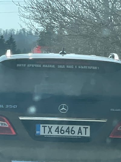 The scene depicts the rear exterior of a dark-colored Mercedes-Benz GL 350 SUV, observed in what is identified as Staro Oriahovo, Bulgaria. The vehicle's tailgate displays a Bulgarian license plate, "BG TX 4646 AT," indicating registration from Varna Province. Above the license plate, the Mercedes-Benz three-pointed star logo is centered, with "GL 350" visible on the lower left and "BLUETEC" partially visible on the lower right of the tailgate. A chrome strip spans horizontally across the tailgate above the license plate. The vehicle's rear window features a white decal with Cyrillic text that reads: "НИТО КРАЧКА НАЗАД. ЗАД НАС Е БЪЛГАРИЯ!", which translates to "NOT A STEP BACK. BEHIND US IS BULGARIA!". The top of the vehicle's roof shows parts of roof rails and a shark fin antenna. The background consists of a natural environment under an overcast sky, with numerous bare tree branches in the foreground, suggesting a late autumn or winter season. Further in the distance, a hillside covered with evergreen trees is visible, and faint red vehicle lights are discernible through the foliage. No individuals are visible.