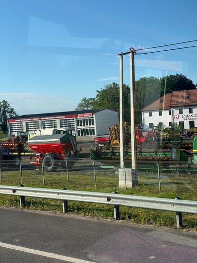The image depicts an outdoor scene, likely a commercial property, with agricultural machinery and buildings. In the foreground, a metal guardrail lines the edge of an asphalt road. Behind the guardrail, a chain-link fence encloses a grassy area with various pieces of agricultural equipment, including a red tractor, a large red seeder with the brand "UNIA" visible, and other harvesting or tilling machinery. In the background, there are two buildings. One is a long, white building with multiple garage doors, labeled in Russian with text that appears to be "СЕРВИС С/Х ТЕХНИКИ • РЕМОНТ ГИДРАВЛИЧЕСКИХ ШЛАНГОВ" (Service of agricultural machinery, repair of hydraulic hoses). Next to it is a white building with a red tile roof and several windows, which has a red circular logo with a white "G" and Russian text that appears to say "ЗАПАСНЫЕ ЧАСТИ НА СЕЛЬХОЗТЕХНИКУ" (Spare parts for agricultural machinery). The sky is blue with some wispy clouds, and trees are visible behind the buildings. There are no people visible in the image. The overall impression is a dealership or service center for agricultural equipment.
