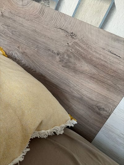 The image shows a close-up view of a wooden surface and a yellow throw pillow. The wooden surface has a grain pattern with knots, and it appears to be a table or desk. The throw pillow is textured and has a fringed edge. The background is blurred but suggests a wall with a pattern. There are no people visible in the image. The scene appears to be indoors. There is no information available to determine the location within the city of Svetlia, Bulgaria.