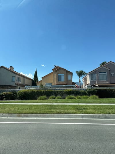 The image shows a view from a street, looking at a row of houses in Fontana, California, USA. The houses are two stories tall with beige and gray stucco exteriors and red tile roofs. They are separated by low walls and hedges. The sky is clear and blue with a few scattered clouds. The foreground consists of a paved road with a white line marking its edge, followed by a strip of green grass and a concrete curb. In the background, some trees and hills are visible.