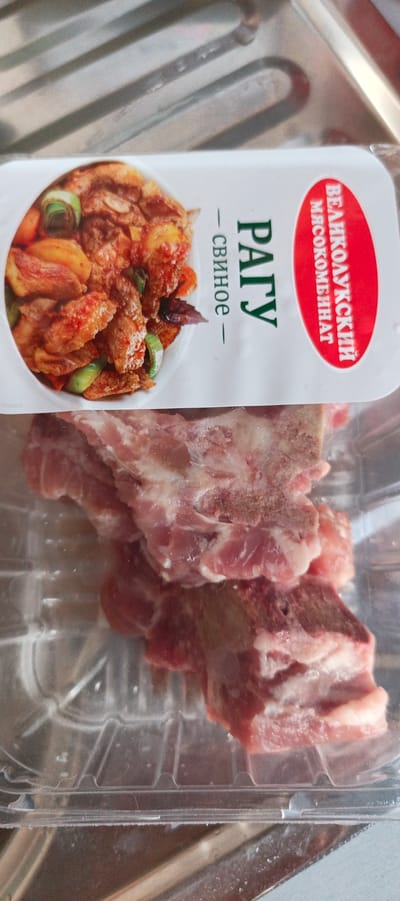 A package of raw pork ribs is displayed on a counter, with a label partially visible. The label features an image of a cooked dish and text in Russian. The ribs are a pale pink to red color with visible white fat and some bone. The packaging is clear plastic. The background appears to be a metallic surface, possibly a kitchen counter or appliance. The lighting is bright, casting some reflections on the packaging and meat. No people are present in the image.