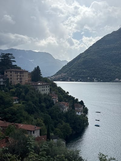 The image displays an elevated view of Lake Como within the city of Nesso, Italy, on an overcast day. The upper portion of the frame is dominated by a sky with extensive light-gray cloud cover, interspersed with areas of brighter sky towards the right. Multiple layers of mountains, predominantly dark green with dense tree cover, frame the scene; a prominent, steeply sloped mountain occupies the right side, descending directly into the lake, while additional hazy mountain ranges recede into the distance on the left and center.
The central and lower-right sections feature a large body of water, Lake Como, exhibiting ripples across its surface and reflecting a silvery-gray light. Three small, unoccupied boats are visible on the water in the middle-right: the top boat is light-colored, the middle boat is dark, and the bottom boat is blue and covered.
The left and central foreground are characterized by a steep, tree-covered incline forming the shoreline, dense with various green deciduous and coniferous trees. Numerous buildings of varying sizes are nestled within this verdant landscape. A prominent, light-colored (beige or cream) multi-story building with multiple window rows stands on an elevated terrace in the mid-left. Below and to its right, another lighter-colored multi-story building with a red-tiled roof is partially visible. Further down the slope towards the lake's edge, several smaller residential structures with terracotta-tiled roofs are interspersed among the trees. On the distant opposite shore, a continuous line of buildings is discernible, extending into the valley between the mountains. No individuals, discernible actions, or interactions are present in the image.