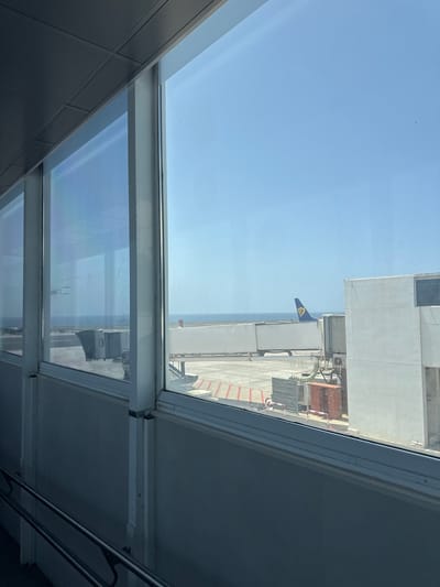 The image depicts a view from inside a building, likely an airport terminal in San Bartolomé, Spain. Through a window, a section of the airport apron can be seen on a sunny day. A blue and yellow flag is visible on a nearby building. The sky is clear and blue. Inside the building, there are visible architectural details such as window frames and interior walls.