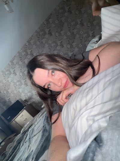 The image is a selfie taken indoors, likely in a bedroom in Башкултаево, Russia. A young woman with long, dark brown hair and light blue eyes is lying on a bed, propped up on her arm. She is wearing a gray t-shirt. Her head is tilted, and she is looking towards the camera with a slight smile. The background shows a wall with floral wallpaper and a glimpse of a dresser and a television. The bed has striped pillows and a gray blanket. The overall lighting suggests it might be evening or a dimly lit room.