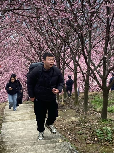 A man in a black puffer jacket and pants stands on concrete stairs. Cherry blossom trees with pink flowers fill the background. Other people are visible in the background, including a couple and a man walking in the distance. The ground is covered in grass and fallen leaves, and the day appears overcast.