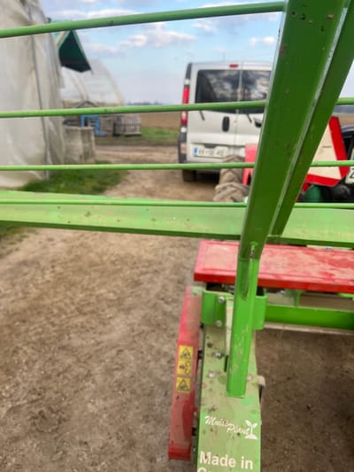 The scene depicts an outdoor agricultural environment in Krško, Slovenia. The immediate foreground features the green painted metal framework and components of a piece of farm machinery, bearing the visible text "Multi Plant" and "Made in" (partially obscured), along with red sections adorned with yellow warning decals. The ground directly beneath this machinery is a light brown, unpaved dirt surface.
In the midground, a dirt path extends backward. To the left of the path, two white polytunnel greenhouses are visible, accompanied by a concrete well and stacked wooden crates. To the right of the path, the rear of a silver panel van, identifiable by a visible Slovenian license plate "KR-3E-492", is parked. Behind the van, a large, lugged tire and a red and white triangular slow-moving vehicle (SMV) sign indicate the presence of a tractor or another piece of agricultural equipment. The background consists of an expansive, uncultivated field under a partially cloudy blue sky. No individuals are present, and no specific actions or interactions are occurring.