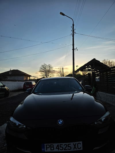 The image shows a black BMW car parked on a street in Krasnovo, Bulgaria. The license plate reads "PB 4525 XC". The car is facing the right side of the frame. The sky is a light blue gradient, suggesting it may be sunset or sunrise. A utility pole with a street lamp is visible on the left side of the frame. In the background, houses and trees are visible. A wooden structure with a roof is also present in the mid-ground. Other vehicles appear to be parked behind the BMW, including a red car.