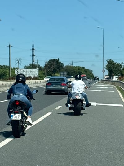 The image shows two individuals riding motorcycles on a sunny day in Constanța, Romania. The motorcycle in the foreground is ridden by a person wearing a blue plaid shirt, blue jeans, a helmet, and white sneakers. The motorcycle has a license plate with the text "CT 41 ALY". In the background, another motorcycle is ridden by a person in a white shirt and blue jeans, also wearing a helmet. They are on a paved road with white lane markings, suggesting a highway or a major road. Trees and power lines are visible on the side of the road, indicating an outdoor, possibly semi-urban or rural setting. The bright sunlight suggests it is daytime, likely afternoon. The overall scene depicts casual motorcycle riding.