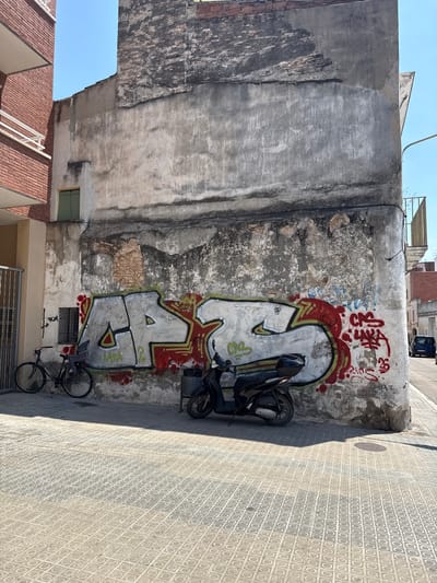 The image shows a graffiti-covered wall in an urban setting, likely Vilanova I La Geltrú, Spain. The wall is old and weathered, with patches of peeling paint and exposed brick. Large, colorful graffiti adorns the wall, featuring abstract shapes and lettering in red, white, and green. A black motorcycle is parked leaning against the wall, partially obscuring the graffiti. A bicycle is also visible, parked on the pavement nearby. The pavement is tiled with a repeating geometric pattern. The scene is bathed in bright sunlight, casting a long shadow across the pavement, suggesting it is daytime. The sky is clear and blue. The overall impression is one of an urban environment with a street art element. The graffiti on the wall appears to include the word "LAKA" in stylized lettering.