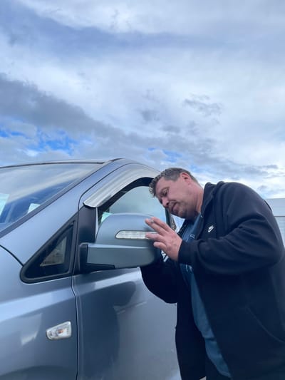 A man is standing outside near a gray car. He is wearing a black hooded jacket and appears to be examining the car's side mirror. The sky is cloudy. The location is likely in a parking area in Бавыкино, Russia, although this cannot be definitively confirmed from the image.