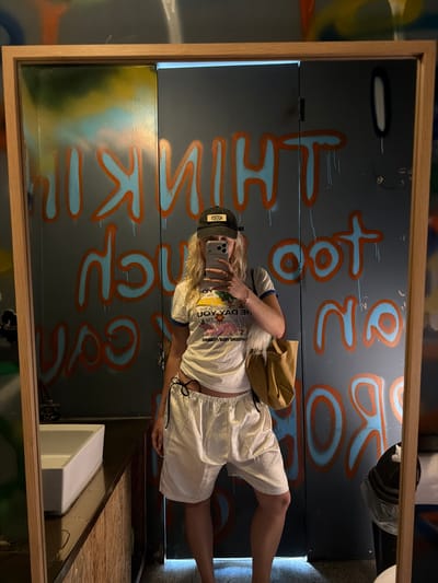 A person stands in front of a mirror in what appears to be a bathroom or restroom. The mirror is framed with light-colored wood, and the reflection shows the individual holding a phone, likely taking a selfie. The person is wearing a white t-shirt, light-colored shorts, and a baseball cap. A tan-colored bag is slung over their shoulder.

Behind the person and reflected in the mirror are gray walls covered with graffiti. The graffiti consists of phrases written in blue and orange paint.

The location appears to be a restroom, as there is a white sink visible in the lower portion of the frame. The sink is partially obscured by a wooden structure.

There is no visual confirmation of the exact location within Toseong-myeon, South Korea.