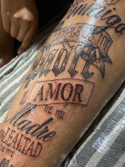 A close-up shows a tan arm covered in fresh, black ink tattoos. Notable words include "Amor," vertically oriented with slight blood around the edges, "Madre," written in cursive, and "Realidad." Other text is partially visible and in a bolder, more ornate font. The tattoos appear to be recent, as they are slightly wet. The arm is resting on a grey and white striped surface.