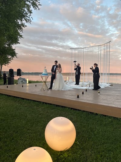 The image captures a wedding ceremony taking place outdoors during a sunset. The location is identified as Osipovo, Russia. In the foreground, a wooden platform extends over a grassy area where two large, illuminated spherical lamps are visible. The platform serves as a stage for the wedding events.

On the stage, a bride and groom stand near a tiered wedding cake placed on an ornate gold table. The groom is wearing a black tuxedo, and the bride is in a white wedding gown. To the right of the couple, two individuals are actively filming the ceremony; one is holding a camera with a gimbal, and the other is operating a video camera. Behind them, a semi-circular backdrop made of hanging white strands creates an ethereal effect.

Further to the left on the stage, a man in a black shirt is standing near audio equipment. In the background, a calm body of water stretches to the horizon, with buildings faintly visible on the opposite shore. The sky above is painted with soft hues of pink, orange, and blue, indicative of a sunset. The overall scene conveys a romantic and celebratory atmosphere.