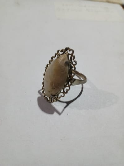 The scene depicts a single antique-style finger ring positioned on a flat, white, or light-colored surface. The location in the city of Phakeo, Laos, cannot be determined from the provided image.

The ring features a large, oval-shaped cabochon stone, light beige to pale cream in color with visible natural inclusions or striations. This stone is set within an ornate, possibly tarnished brass or gold-colored metal bezel with a filigree design comprising scrolled elements. The ring's band is a silver-colored metal, exhibiting some wear and minor surface imperfections. A faint "900" marking is visible on the exterior of the band.

The object is illuminated by a directional light source from the upper left, casting a distinct, dark shadow of the ring towards the lower right. The background consists of the same light-colored surface, out of focus, with subtle texture and faint horizontal lines in the upper portion. No individuals, actions, or interactions are present.