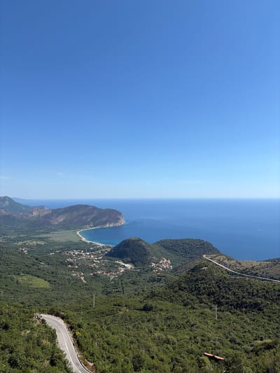 The image presents a panoramic view from a high vantage point overlooking a coastal region. In the foreground, a winding paved road cuts through lush green, densely forested hills. The road features white dividing lines and appears to be in good condition. Further into the middle ground, the landscape transitions to a flatter area with scattered buildings, forming a small village. Beyond the village, a crescent-shaped sandy beach lines the azure blue sea. The sea stretches to the horizon, meeting a clear, bright blue sky with no clouds visible. In the distance, mountainous terrain frames the coastline. The scene appears to be devoid of people. This view is consistent with overlooks found in the vicinity of Novoselje, Montenegro, a location known for its scenic coastal and mountainous landscapes.