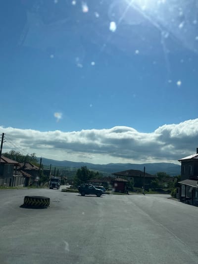 The image depicts a street scene in Rish, Bulgaria. The sky is predominantly blue with scattered clouds. A line of puffy, elongated clouds stretches horizontally across the midground. The foreground is a gray asphalt road, with a large tractor tire lying on the side. Several vehicles are parked along the side of the road, including a dark-colored car and a white truck. Buildings are visible on either side of the road, with red tile roofs and gray facades. Mountains can be seen in the distance behind the buildings. Power lines are also visible. The overall perspective suggests the image was taken from within a vehicle.