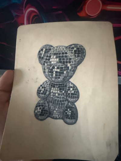 The image shows a close-up of a paper with a drawing of a disco ball teddy bear. The drawing is rendered in a grayscale, pixelated style, mimicking the reflective surfaces of a disco ball. The teddy bear drawing is centered on the paper. The paper is a light beige color and appears to be slightly worn or aged. The background of the image is out of focus and shows abstract shapes and colors, possibly indicating the paper is lying on a desk or table with a colorful item behind it. A hand is partially visible on the left side of the image, holding the paper. There is no indication of a specific location in the city of Leominster, United Kingdom, or any other city. The scene is static and focuses solely on the drawing and the paper.