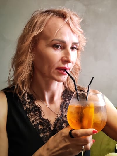 A medium shot captures a Caucasian woman with blonde hair, wearing a black dress with lace trim, holding a large glass of an orange beverage with two black straws. She is shown from the chest up, with her head tilted and her gaze directed downwards as she sips the drink. Her right hand is visible, adorned with red nail polish and two rings, one black and one silver. The background is a neutral, textured gray wall. The scene is set in Chelganovo, Russia.