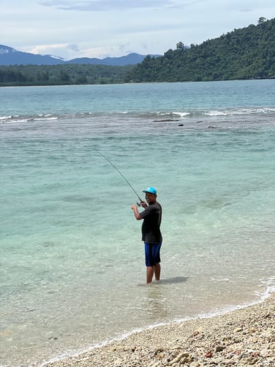 In the city of Pasi, Indonesia, a person stands in the shallow, clear turquoise water near a pebble beach, fishing with a rod and reel. The person is wearing a black t-shirt, blue shorts, and a blue baseball cap. The person is looking towards the camera with a smile and holding the fishing rod. In the background, the calm sea extends to a distant treed coastline, with misty mountains beyond. The sky is cloudy.