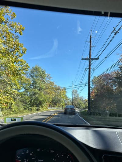 The image displays a forward-facing perspective from inside a vehicle driving on a two-lane asphalt road in Lawrence Township, New Jersey, United States. The sky is clear blue, indicating daytime conditions. The road features a double yellow center line, a white shoulder line on the right, and curves to the right in the mid-distance. Trees with predominantly green and yellow autumn foliage line both sides of the road. A dark-colored sedan is visible ahead, traveling in the same direction on the right side of the road. Multiple wooden utility poles, heavily laden with electrical or communication lines, are positioned along the right side of the road. A white guardrail is visible on the left side of the road as it curves. The bottom of the image shows the interior of the observer's vehicle, including part of the steering wheel with a grey rim and the dashboard displaying gauges. A digital readout on the dashboard indicates 66°F and 1:53 PM. A green and white New Jersey temporary motor vehicle inspection sticker is affixed to the lower-left portion of the windshield.