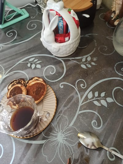 The image displays a close-up, overhead view of a table setting, likely a breakfast or snack. On a dark, patterned tablecloth, a white, ceramic bunny-shaped dish holds a red and blue packaged item with visible Russian text. To the right, a plate with a decorative gold rim holds several small, golden-brown pancakes and a clear glass teacup filled with a dark liquid, presumably tea or coffee. A silver spoon rests on the tablecloth to the upper right. A light blue stand is visible on the lower left, holding what appears to be a smartphone. The scene is well-lit, suggesting it's daytime. The overall impression is one of a simple, home-based meal or break. The text on the packaging is partially visible and reads "ДЕСЕРТ ГЛАЗИРОВАННЫЙ" and "ЛОМТИШ". Verkhnii Lars, Russia is the provided location context, implying this scene is set there.