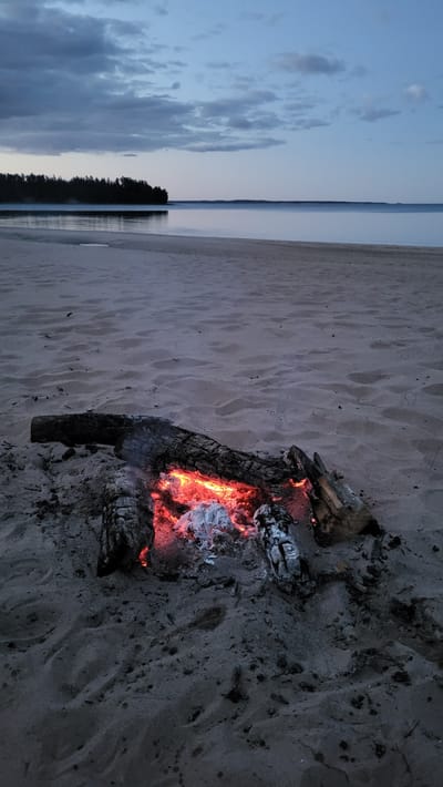 A low-angle, full outdoor shot captures a campfire on a sandy beach in Zaostrov'e, Russia. The fire, in the foreground, consists of charred logs and glowing embers. Behind the campfire, the scene transitions to a vast, calm body of water that meets a distant, dark treeline under a twilight sky with scattered clouds. The sand is textured with footprints and small debris, suggesting a natural, unpopulated setting. The lighting indicates it is either dusk or dawn.