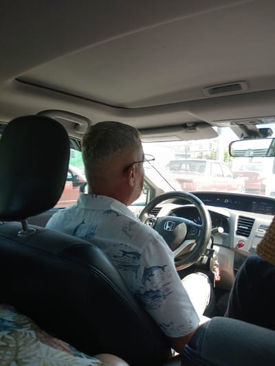 The image shows the interior of a car, with a man driving. The man is wearing glasses and a short-sleeved shirt with a dolphin pattern. He has short, gray hair. The car's dashboard is visible, including the steering wheel, instrument cluster, and center console. Outside the car, other vehicles and buildings can be seen, suggesting an urban environment. The time stamp on the dashboard indicates 15:52. The car appears to be on a street in the city of Karatay, Turkey, as evidenced by the license plate on the red car in front of the driver, which reads "42 ECR 647". The "42" is the province code for Konya, the larger administrative region that Karatay is part of.