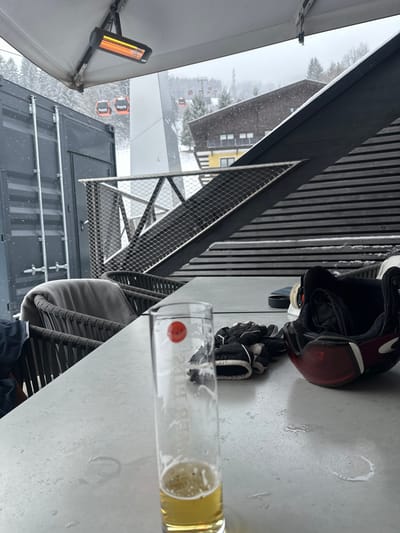 The image presents a forensic view of an outdoor terrace in Bad Hofgastein, Austria, during light snowfall. In the immediate foreground, a light-colored rectangular table holds a tall, clear pilsner glass containing approximately one inch of yellow liquid (beer) and a small amount of foam, with a red circular logo and faint text visible on its side. To the right of the glass, a pair of black and white ski gloves are folded, and a red and black ski helmet with goggles attached rests on its side. Water droplets and faint ring marks are visible on the table surface.
To the left of the table, a dark wicker-style chair is partially visible, draped with a grey blanket. Behind it, a dark grey shipping container-like structure with vertical ridges and a ladder extends upwards. Overhead, a black infrared patio heater with an active orange glow is mounted beneath a grey awning or roof structure. A metal railing with a diamond-patterned mesh infill runs diagonally across the mid-ground, separating the terrace from the vista. Beyond the railing, a dark, angular structural element with horizontal slats is observed.
The background reveals a snow-covered mountainous landscape dotted with pine trees. Multiple orange gondola cabins, each bearing the "Gastein" logo, are visible on a ski lift line ascending a slope. Several buildings, including one with a dark brown roof and a yellow lower facade, are nestled among the trees. Snowflakes are discernible in the air, indicating active precipitation.