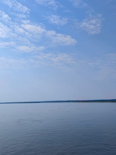 The image depicts a wide, calm body of water, likely a river or lake, under a bright sky filled with scattered white clouds. The horizon line is clearly visible, separating the water from a distant, flat shoreline covered in green vegetation. The water's surface is smooth with subtle ripples, reflecting the light of the sky. The overall scene conveys a peaceful and expansive natural environment. No people or specific man-made structures are discernible in the foreground or mid-ground of the image, apart from faint outlines of what might be structures or industrial elements along the distant shoreline, barely visible against the horizon. The water appears to be a deep blue-grey color, and the sky transitions from a pale blue near the horizon to a slightly deeper blue overhead. The light suggests it is daytime, possibly late morning or early afternoon, with the sun positioned high in the sky.