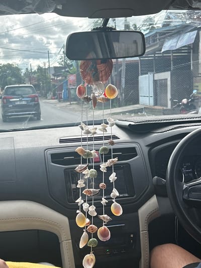 The image depicts the interior of a car, viewed from the driver's perspective, looking out through the windshield. A seashell wind chime hangs from the rearview mirror, its various shells dangling in the foreground. Outside the car, a street scene unfolds, showing a dark-colored SUV in front, a road, and buildings lining the street. The sky is overcast with clouds. The dashboard of the car is visible, including the steering wheel, instrument cluster displaying "228" and "82 192", and a car stereo. The car's interior features light-colored upholstery on the passenger side. The exterior scene appears to be in A R A, Indonesia, given the presence of Indonesian license plates on the vehicle and the style of buildings.