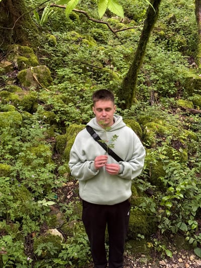 A man is standing in a lush, green environment that appears to be a forest or heavily vegetated area. The man is wearing a light green hoodie, black pants, and a black shoulder strap. He is holding a small leafy branch. The ground is covered in moss, various plants, and small rocks. The environment appears natural and somewhat wild. There are no visible man-made structures, suggesting the image was taken outside of urban Tsabal, Abkhazia, in a heavily wooded area or surrounding nature.