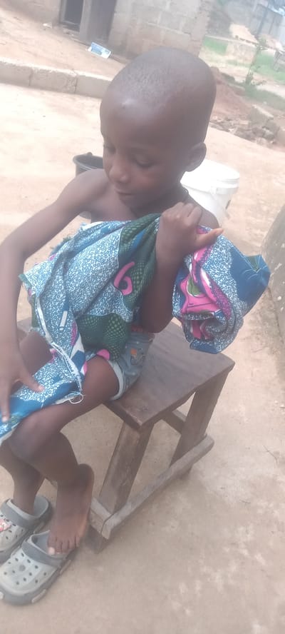 A young child sits on a wooden stool outdoors in Bende, Nigeria. The child is light-skinned, with a shaven head and is draped with a colorful patterned cloth. He wears Crocs-like sandals and what appears to be shorts with bicycle designs. The background features a concrete building, a white bucket, and a dirt ground.