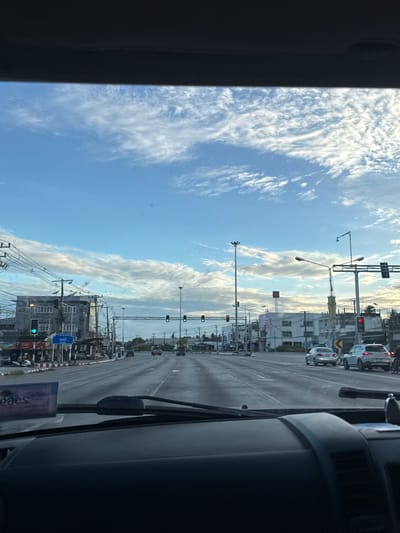 The image shows a view from inside a vehicle looking out at a street scene in Ban Nong Mu, Thailand. The sky is partly cloudy with streaks of white clouds against a blue sky. The road is wet, suggesting it has recently rained or is about to. Several vehicles, including cars and motorcycles, are visible on the road. Traffic lights are present, with some showing green lights. Buildings line the sides of the road, appearing to be commercial establishments. Power lines and streetlights are also prominent features of the urban landscape. The overall impression is that of a typical street scene in a Thai city, likely during the daytime.