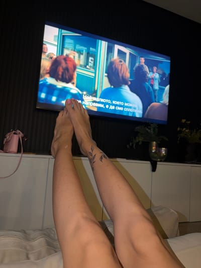 A forensic examination of the image reveals an indoor environment, likely a residential space, characterized by subdued lighting. In the foreground, a pair of light-skinned human legs and feet are elevated and extended, resting on a light-colored, possibly textile, surface. The toenails on both feet are painted a pale pink. A small, off-white adhesive bandage is visible on the inner aspect of the left ankle. The left calf displays a tattoo featuring a black ink vine pattern with leaves.

In the mid-ground, a large, rectangular flat-screen television is mounted on a dark, vertically textured wall. The television screen is active, displaying a scene that includes several individuals, primarily viewed from behind, within what appears to be an outdoor setting featuring a light blue building structure. A man in a blue shirt is more clearly visible in the background, facing the camera. White Cyrillic subtitles are present at the bottom of the screen, reading: "Най-малкото, което можем да направим, е да сме сплотени." (The least we can do is be united.)

Below the television, a long, low white cabinet unit with visible seams suggesting modular compartments is situated. On the left end of the cabinet, a small, pale pink crossbody bag with a slender chain strap is positioned. Towards the right, a potted plant with green foliage is contained within a clear, possibly iridescent, glass vessel. Further right, a dark pot containing yellow flowers is partially visible.

The specific city location in Tassoultante, Morocco, cannot be determined from the visual information provided within the image.