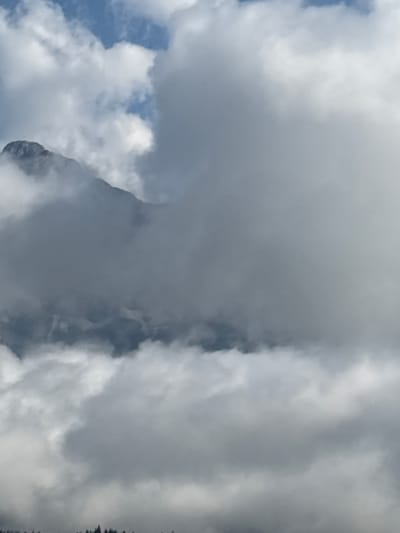 The image captures a mountainous landscape in or near Kleinvolderberg, Austria, with a prominent peak partially obscured by heavy clouds. The sky is a mix of blue and white, with dense, fluffy clouds dominating the scene. The mountain itself appears rugged and dark, its upper sections shrouded in mist. The lower portion of the image shows a dark, indistinct treeline, suggesting the presence of a forest. There are no people or identifiable objects present, creating a natural and somewhat dramatic atmospheric depiction of the mountain environment.