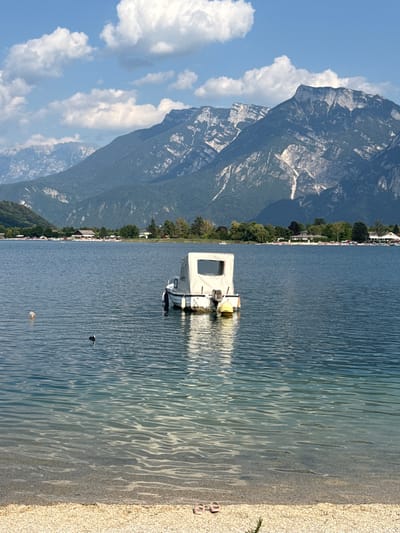 This is a wide shot photograph of a lake with a boat docked on the water, set against a backdrop of mountains. The water is calm and reflects the blue sky and clouds. In the foreground, there is a small, pebbly beach with a pair of pink sandals. In the middle ground, a white boat with a covered cabin is moored. There are a few buoys in the water around the boat. Further in the distance, a shoreline is visible with trees, buildings, and a beach with people. The background is dominated by large, imposing mountains with rocky, uneven surfaces, some covered in vegetation. The sky is blue with scattered white clouds. The scene depicts a peaceful day at a lake, likely Lake Caldonazzo, in or near the town of Calceranica al Lago, Italy, given the presence of mountains and a lake commonly associated with the region.