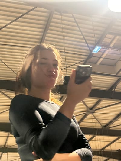 A woman in a dark long-sleeved shirt holds a phone upward, presumably taking a photo or video. She stands under a large structure, possibly a building or stadium, with a ceiling of bright white corrugated metal panels and steel beams. A rectangular light fixture is visible on the ceiling. Her hair is light brown and her face is illuminated, suggesting an upward gaze.