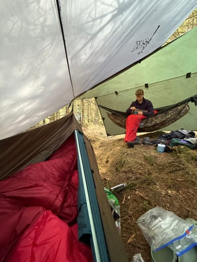 The image shows a campsite in Macclesfield Forest, United Kingdom, with a person lying in a hammock under a tarp. The hammock is hung between two trees and the tarp is attached to the trees. There is a red sleeping bag in the hammock. The person is wearing a dark jacket and red pants and is eating from a mug. To the left of the hammock is a closed tent, with a red sleeping bag inside. The ground around the campsite is covered in brown leaves, and there are some objects scattered around the site. The photo is taken from a low angle looking up at the tarp and the person in the hammock. The location of this campsite is 53.26 latitude and -1.991 longitude, a forested area with towering trees surrounding the site.