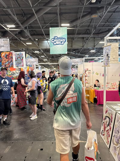 This image depicts the interior of the Paris-Nord Villepinte Exhibition Centre in Villepinte, France, during an event, likely a convention or expo, given the presence of booths and displays. The scene is bustling with activity, with numerous people browsing and interacting within the venue.
In the foreground, a person wearing a grey baseball cap and a green and white striped jersey walks away from the camera, carrying a white shopping bag. The jersey features a dragon graphic and Japanese characters. The person's legs and lower torso are visible, clad in light-colored shorts and black sneakers.
Behind this individual, the event space opens up, revealing several exhibition booths. To the left, a wall displays vibrant artwork, including a fiery demon-like character and a traditional Japanese mask. Several people are seen in this area, including a person in a blue t-shirt with the Paris 2024 Olympics logo, a woman in a red plaid dress, and others browsing the artwork.
Further into the venue, more booths are visible, with tables laden with merchandise. In the background, banners hang from the ceiling, one prominently displaying "CREATORS WORLD" in large, stylized text. The architecture of the exhibition center is evident, with exposed metal structures, ductwork, and lighting fixtures on the ceiling. The floor is a dark, reflective surface, marked with lines and some scuffing.
The overall atmosphere suggests a lively and engaging event, with a focus on art, commerce, and popular culture.