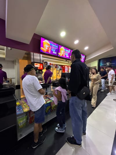 The image depicts an indoor scene at the concession stand of Teatro Cecilia, a cinema located in Pitalito, Colombia. In the immediate foreground, a man wearing a white t-shirt, blue shorts, and a black baseball cap (bearing "VR46") stands facing right, positioned in front of a glass display case containing packaged snack foods such as Doritos and Todito. To his right, a taller man, observed from the back, wears a black jacket, blue jeans, and carries a black messenger bag across his body; he stands alongside a young girl in dark pants and a pink top or wrap. All three individuals are oriented towards the concession counter.

Behind the counter, two cinema staff members in purple uniforms are visible, one operating a Coca-Cola soda dispenser on the left, and another positioned further right near what appears to be a popcorn machine. Above the counter, three illuminated digital screens display "COMBO MAX" advertisements featuring images of popcorn, soft drinks, and hot dogs, with associated pricing in Colombian Pesos, such as "$47.900." To the right of the central group, a woman in a light brown two-piece outfit (crop top and flared pants) stands facing left. Additional patrons, predominantly male, are visible in the background to the right, forming a line or gathering. The floor consists of black and white tiles, while the walls are colored purple and off-white. The ceiling is white with recessed lighting. A security camera is mounted on the ceiling in the upper left quadrant of the frame.