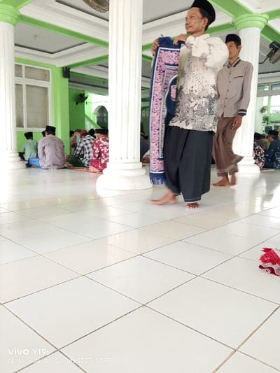 Inside a mosque, several Muslim men are present, some seated on the floor in prayer while others are standing. One man, wearing a white patterned shirt, black sarong, and a black peci hat, is holding a prayer rug. Another man in a light brown shirt and brown trousers is walking alongside him. The room features white tiled flooring and green pillars with white bases. A red and white cloth is visible on the floor in the foreground.