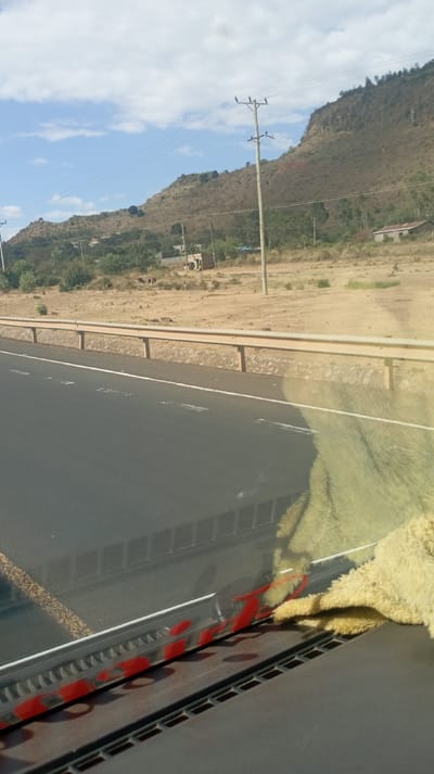 A road with a metal guardrail runs parallel to a dry, scrubby landscape. A low, rugged hill rises in the background, punctuated by scattered trees and utility poles. Several buildings are visible in the distance. The foreground contains a dashboard with a suction cup mount and a furry object, likely a stuffed animal. The scene is taken from inside a vehicle, looking out through the windshield.