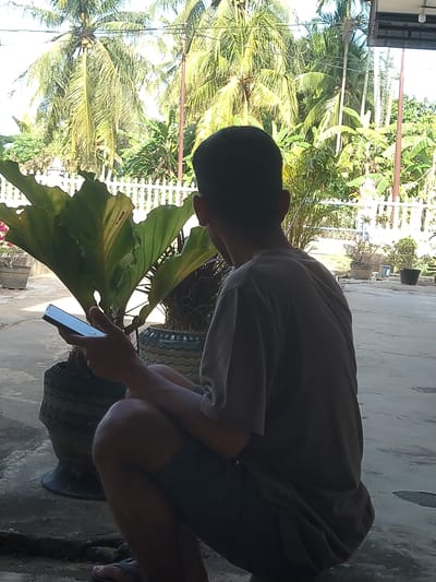 A low-angle, portrait-oriented photograph depicts a person crouched outdoors, interacting with a potted plant while holding a smartphone. The image is presented on its side, with the ground on the right and the sky on the left.

The central subject is an individual, likely male, with short dark hair, wearing a dark grey t-shirt and dark shorts. He is shown from a side profile, with his back slightly turned, in a crouched or kneeling position. His face is mostly obscured in shadow as he looks downwards with focus. In his right hand, he holds a dark-colored smartphone, positioning it near the base of a large, leafy green plant. This suggests he might be photographing the plant or using the phone for related information.

The plant is a prominent feature, showcasing broad, dark green leaves, possibly belonging to a tropical species like Anthurium. It is potted in a dark, textured container, which appears to be a repurposed tire. The person's left hand is also near the plant's foliage.

The background reveals a vibrant, tropical landscape, dominated by numerous tall palm trees with their distinctive fronds. Other dense green foliage and what might be a fence or railing are also visible. The ground is a light-colored, paved surface, suggesting an outdoor area such as a patio or courtyard.

The lighting indicates a bright, sunny daytime, with strong sunlight illuminating the background vegetation and casting some shadows. The person's front is in shadow, creating a subtle silhouette against the brighter background. No visible text is present in the image. The abundance of tropical plants, particularly palm trees, aligns with the provided location context of Gampongbaro, Indonesia, indicating a warm, verdant environment.