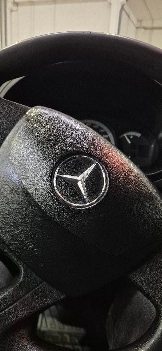 This image depicts the interior of a vehicle, specifically focusing on the black steering wheel, located in Santa Perpètua de Mogoda, Spain. The central airbag cover of the steering wheel prominently features a chrome Mercedes-Benz three-pointed star logo encircled by a chrome ring, set against a textured black surface. Portions of the vehicle's instrument cluster are visible, out of focus, behind the steering wheel, showing indistinct gauges and indicators. The wider background suggests an indoor environment, possibly a garage or service bay, with light-colored walls or ceilings and overhead lighting visible, all blurred. No individuals are present, and no specific actions or interactions are depicted.