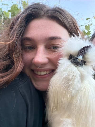 A young woman with brown hair and freckles is holding a fluffy white chicken to her face. She is smiling, and the chicken's head is partially obscuring her face. The background is blurred, but appears to be outdoors with some greenery and a light blue sky. It is not possible to determine the location in Uhersko, Ukraine from the image.