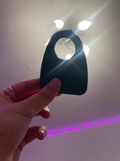 A hand, wearing a silver ring on the index finger, holds a dark, tear-drop-shaped object with a circular cutout. The object is positioned against a ceiling with visible light fixtures emitting bright light. Below the hand, a string of pink LED lights is visible, creating a gradient of pink and purple light. The background appears to be an indoor setting. There is no indication of the location being Vaganovo, Russia.
